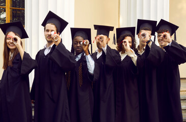 Portrait of happy graduate students standing in a row and looking through diploma like a telescope...