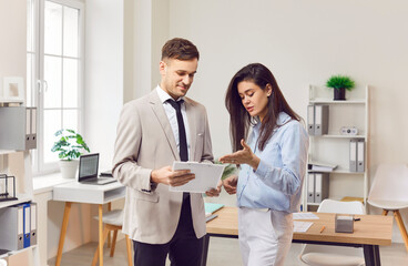Portrait of two smiling business people man and woman standing in office at their workplace and looking through financial documents. Employees discussing work project or company finances.
