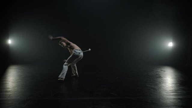 Group of a professional dancers are practicing capoeira in darkness against a spotlight on a black background of studio. Afro-Brazilian martial art that combine elements of dance.