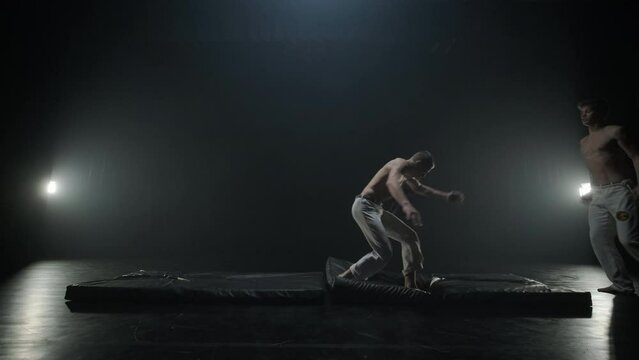 Group of a professional dancers are practicing capoeira in darkness against a spotlight on a black background of studio. Afro-Brazilian martial art that combine elements of dance.