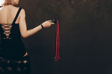 Slender Woman holding Black and Red Leather Flogging Whip in hand in dark interior, view from back