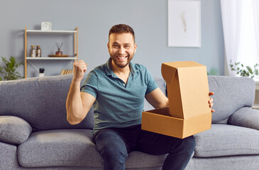 Happy man sitting on sofa and open a cardboard box at home, receiving a parcel, package or gift....