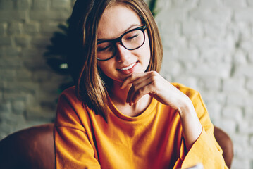 Positive hipster girl in trendy eyeglasses resting in cafe interior chatting in social networks via...