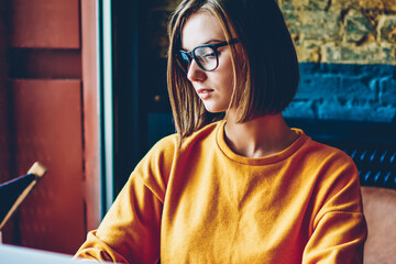 Smart concentrated female copywriter in eyeglasses working freelance at laptop device connected to...