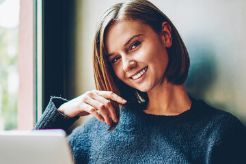 Portrait of pretty hipster girl smiling at camera while resting on windowsill in coworking with...