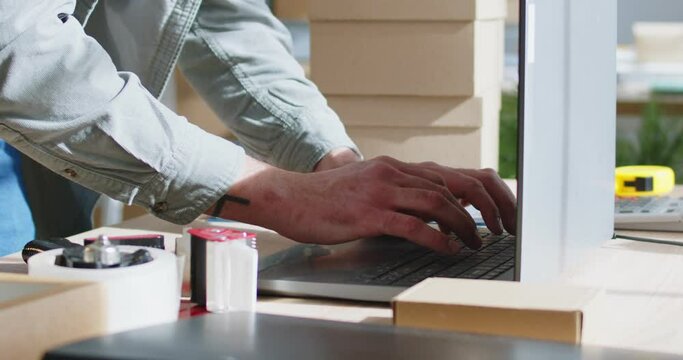 Male warehouse worker efficiently processes online orders using laptop at workplace. Close up of man's hand typing on laptop standing on table next to adhesive tape, tape measure and cardboard boxes. 