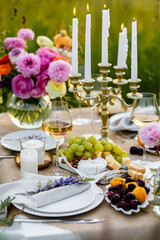 Romantic sunset wedding table decor for a couple with vintage chandelier, burning candles, snacks, white wine, fruits, ranunculi flowers. Summer vibes, field or meadow, outdoors event