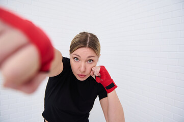 Determined female fighter with red tapes on her fists, punching forward looking at camera, isolated over white wall background. European young woman boxer 35-40 s practicing sport, boxing training
