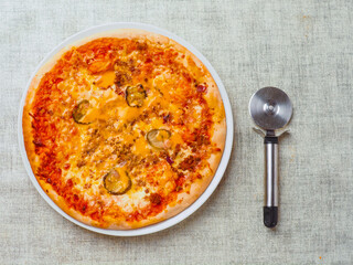 A pizza with cheese and pickles on top and shiny metal pizza cutter. Italian dish with dough, sauce and cheese. Simple white table cloth. Popular food.