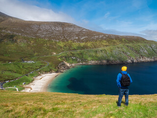 A man in a blue jacket stands on a hill overlooking a beach and a ocean. The sky is clear and the sun is shining, creating a peaceful and serene atmosphere. Keem bay and beach Ireland. Travel concept