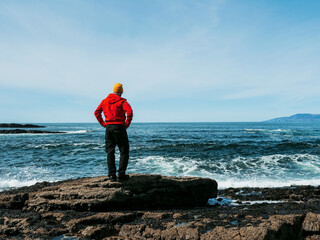 A man in a red jacket stands on a rock overlooking the ocean. The man is wearing a yellow hat and he is looking out at the water. The scene is peaceful and serene. West coast of Ireland. Travel theme