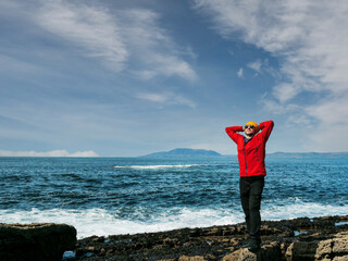 A man in a red jacket stands on a rock overlooking the ocean. The man is wearing a yellow hat. The...