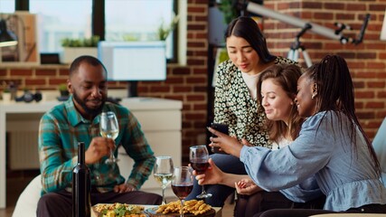 Multiracial friends at apartment party looking at social media posts made by acquaintance on...