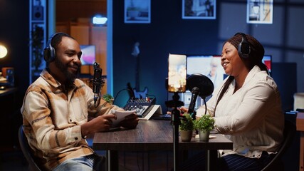 Vlogger show host and guest using smartphone on tripod to record discussion in neon lights ornate personal studio. Man and woman filming online podcast episode with mobile phone