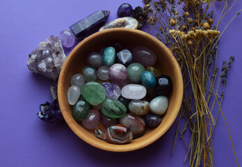 Beautiful natural semi-precious stones in a wooden plate and dry herbs on a purple background. Predictions, witchcraft, spiritual esoteric practice.
