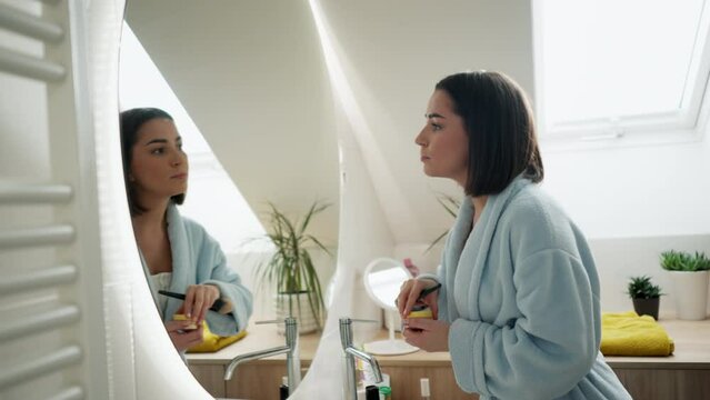 Attractive young woman doing daily makeup while standing near mirror in bathroom