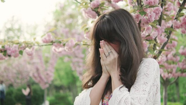 Sick brunette woman having runny nose and suffering from seasonal allergy near blooming sakura tree