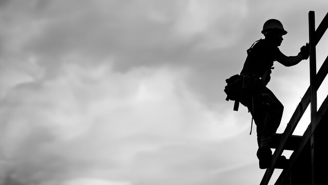 Silhouette of a construction worker at top structure wearing a hard hat in black and white with high contrast against a sky background