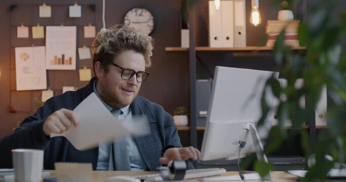 Loop of tired businessman working with computer typing and waving papers feeling hot sitting at table in office in evening. People and workplace concept.