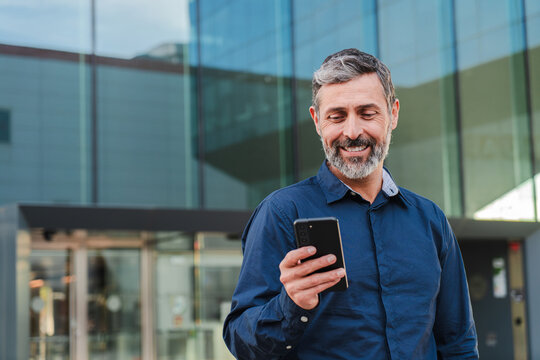 Real mature male worker watching a finance app on his cellphone. Middle aged person holding a mobile phone smiling. Handsome mid adult man smiling using a smartphone at workplace.