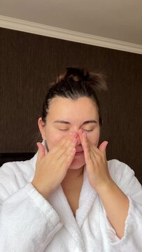 Cosmetic procedure. Young adult woman in white bath robe applies moisturizer to face in hotel bed room. Vertical video. Concept of march 8, rest, skin protection during cold season, spa salon, relax