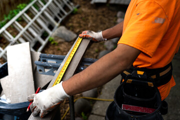 Construction worker in an orange shirt using a tape measure and pencil to mark wood for cutting.
