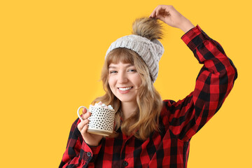 Pretty young woman holding cup of hot chocolate with marshmallows on yellow background
