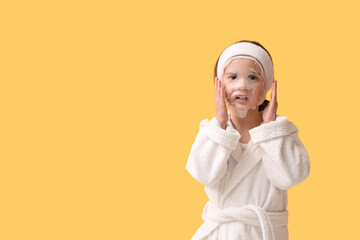 Cute little happy girl in bathrobe with sheet facial mask on yellow background