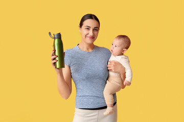 Beautiful young sporty woman with bottle of water holding her little baby on yellow background