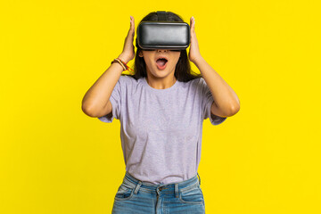 Excited happy Indian young woman using headset helmet app to play simulation game. Watching virtual reality 3D 360 video. Arabian girl in VR goggles isolated on yellow background. Future technology