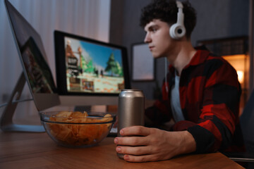 Young man with energy drink and headphones playing video game at wooden desk indoors, focus on can