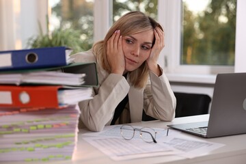 Overwhelmed woman sitting at table with stack of documents and folders in office