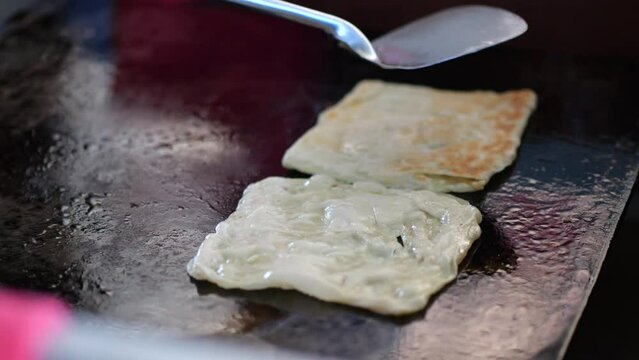 Street food vendor is preparing the dough and pan-frying paratha roti in hot oil until crispy on the outside. Street hawker preparing prata bread or roti canai for morning breakfast