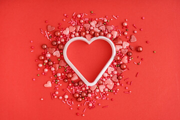 Heart shaped cookie cutter with sweet colorful sprinkles scattered on red background