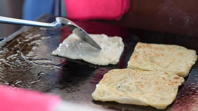 Street food vendor is preparing the dough and pan-frying paratha roti in hot oil until crispy on the outside. Street hawker preparing prata bread or roti canai for morning breakfast