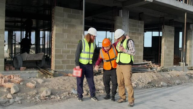 Injury on construction site, worker injured leg in fall, walks with the support of medical staff holding a first aid kit and a smartphone to receive emergency care.