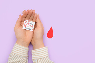Female hands holding paper piece of sheet with text WORLD BLOOD DONOR DAY and paper drop on lilac background