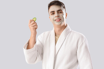 Young man with applied clay mask and cucumber slice on light background