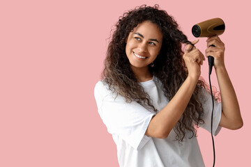 Beautiful young happy African-American woman blow drying her healthy curly hair on pink background
