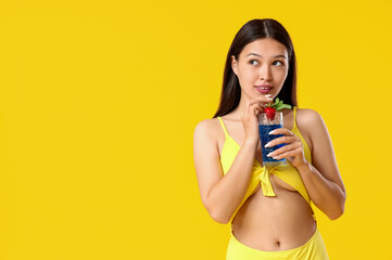 Beautiful young Asian woman in swimsuit drinking cocktail on yellow background