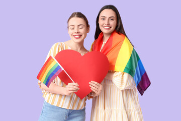 Beautiful young lesbian couple with LGBT flags and paper heart on lilac background