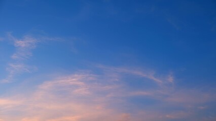 A blue sky transitioning into the soft colors of sunset. Wispy clouds are tinted with light pink and orange hues, creating a calm and serene evening atmosphere. Sunset sky background.
