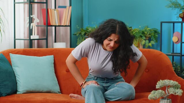 Sick ill Indian woman suffering from period cramps, painful stomach ache on sofa at home room. Girl holding belly, feeling abdominal menstrual pain. Abdominal pain, gastritis, diarrhea, indigestion