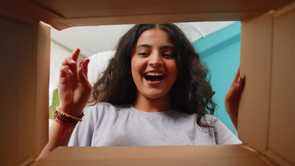 View from inside the box. Happy Indian woman unpacking delivery parcel at home. Smiling satisfied girl shopper, online shop customer opens cardboard box receiving purchase gift by fast postal shipping