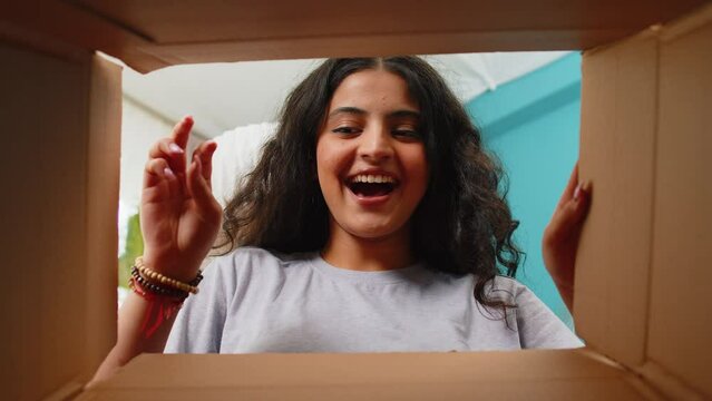 View from inside the box. Happy Indian woman unpacking delivery parcel at home. Smiling satisfied girl shopper, online shop customer opens cardboard box receiving purchase gift by fast postal shipping