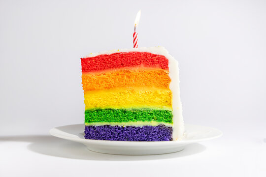 A slice of cake made of a rainbow of colors upright with a candle on a round white plate on a white background