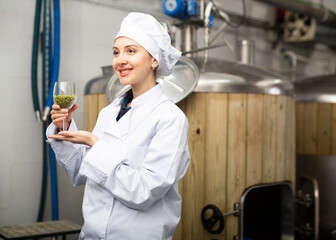 Portrait of positive woman brewmaster holding glass of hops pellets in beer factory.