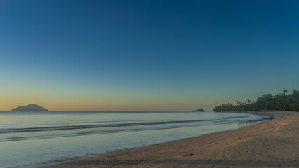 Evening seascape. The waves of the ocean are calmly rolling towards the shore. Shadows on the sandy beach. Green trees, an island in the distance.The blue sky is highlighted with orange at the horizon