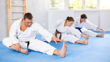 Obraz premium Male and female visitors of martial arts class are preparing for lesson. Teacher and followers perform warm-up exercises and legs stretching. Preparation for sports event