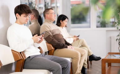 Guy browsing websites on smartphone while sitting in lobby of job center or employment office, waiting for interview or conversation with career counselor